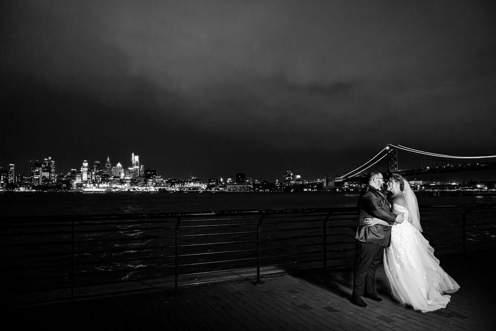 Night time photo with bride and groom posing with the City of Philadelphia skyline in the background at the Adventure Aquarium wedding in Camden NJ