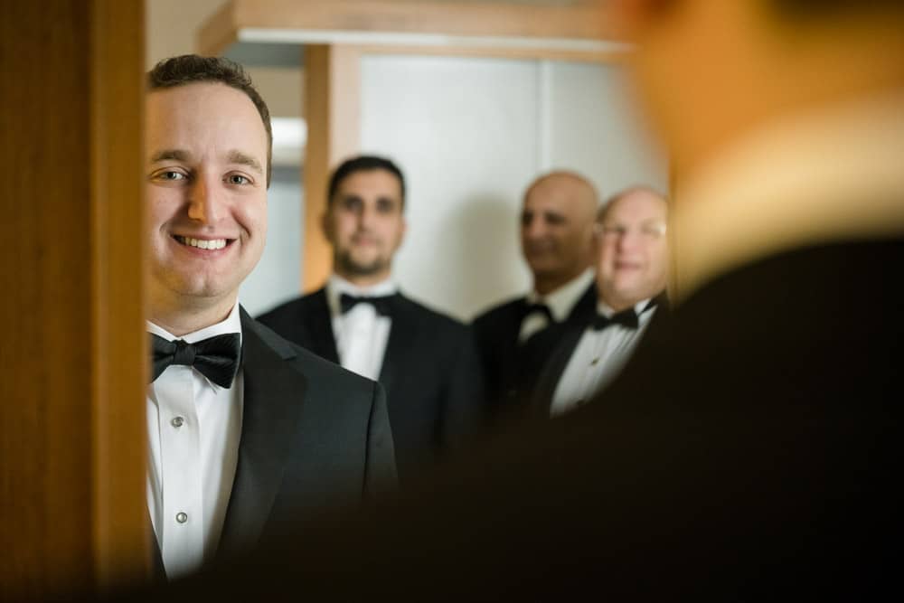 Groom looking at his reflection in the mirror with groomsmen in the background at the Legacy Club at Woodcrest in Cherry Hill NJ