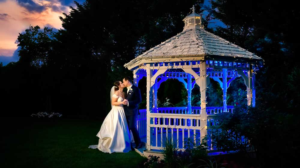 Beautiful sunset with the groom and bride standing next the gazebo lit in a deep blue color at Bradford Estate.