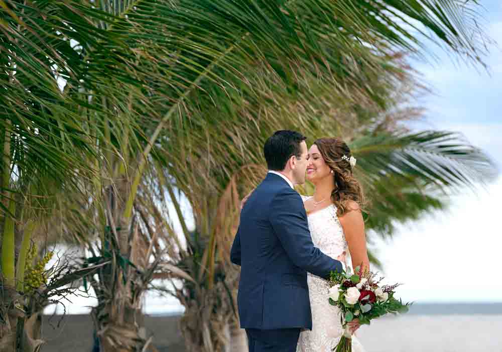 Bride and groom embrace on the beach by the palm trees at the ICONA Diamond Beach wedding.