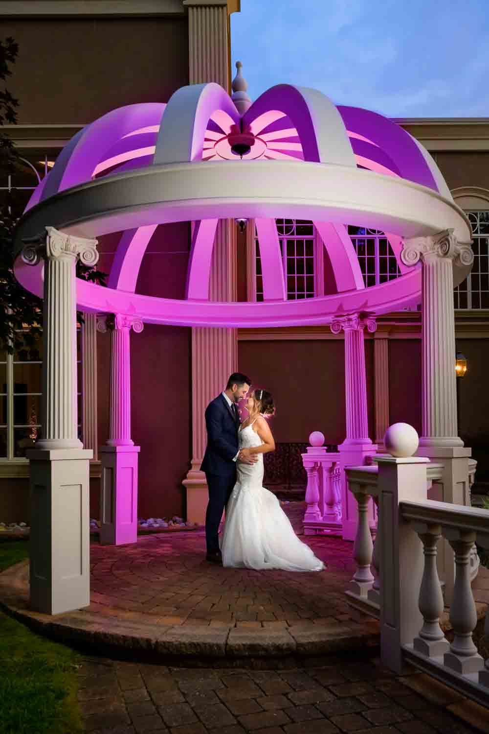 Michael and Laura posing inside the stone gazebo that is beautifully lit in Magenta by the photographer at Brigalias wedding.